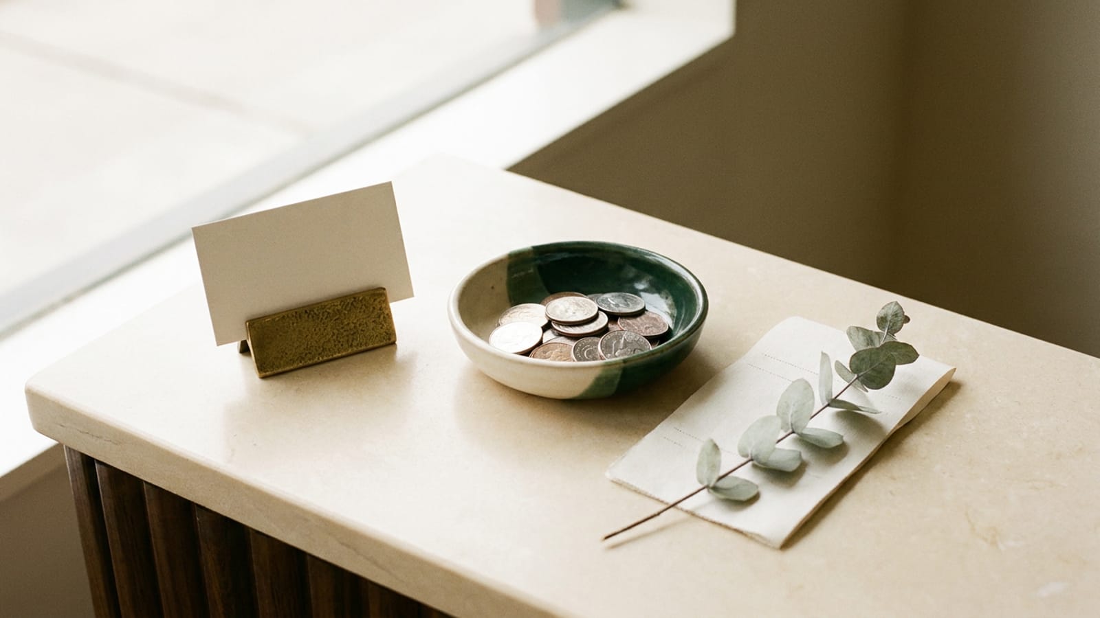 Comptoir de salon vu de haut : un petit chevalet porte-carte vide, une coupelle en céramique avec des pièces et une branche d’eucalyptus séchée